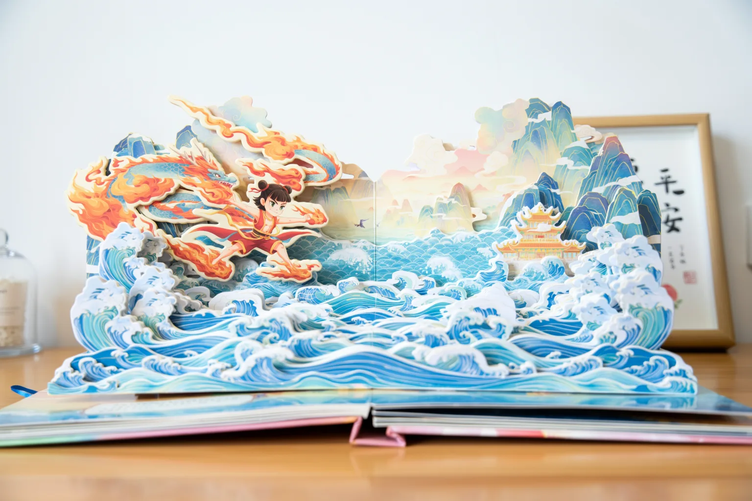 A paper pop-up book open on a desk showing the Nezha Conquers the Dragon King scene in layered paper waves